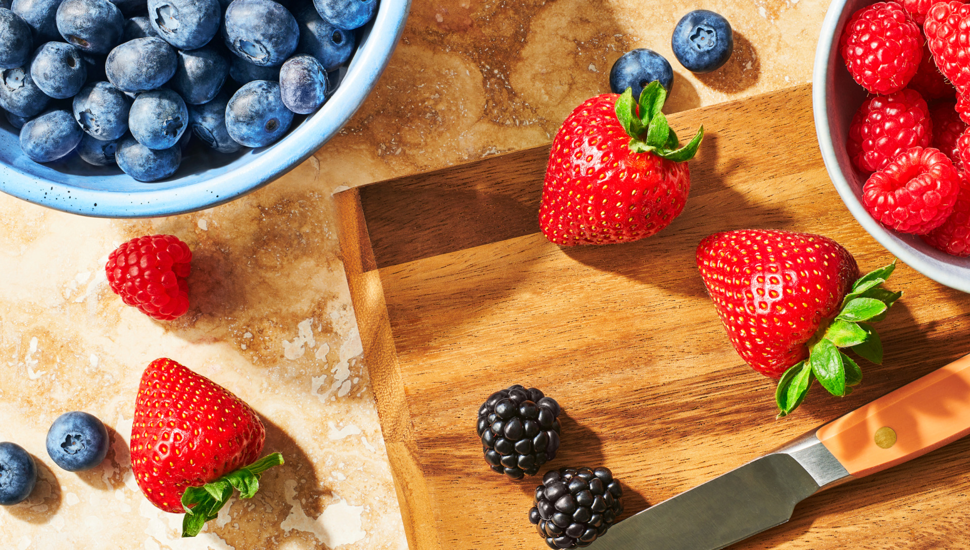 Image of a bowl of Always Fresh blueberries, a bowl of Always Fresh raspberries with strawberries, blackberries, raspberries and blueberries on a cutting board with a knife.